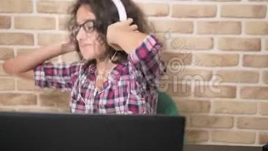酷酷的少年男孩，一头卷曲的黑发，穿着格子衬衫，戴着耳机，跳舞，坐在笔记本电脑前